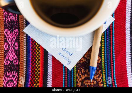 Tazza di caffè con una nota che dice Buongiorno su un tavolo colorato con intreccio Inca. Foto Stock