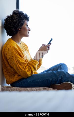 Donna afroamericana seduta su una tenera finestra con lo smartphone e guardando all'esterno, spazio per copiare Foto Stock