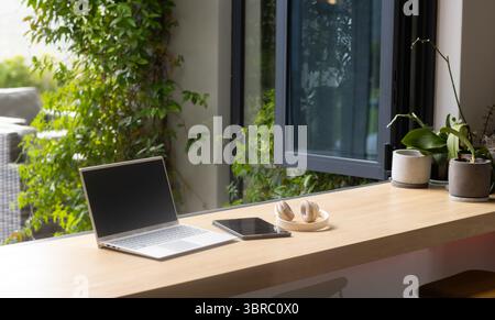 Computer portatile in argento aperto con tablet e cuffie su un bancone in legno vicino a una finestra aperta, con piante Foto Stock