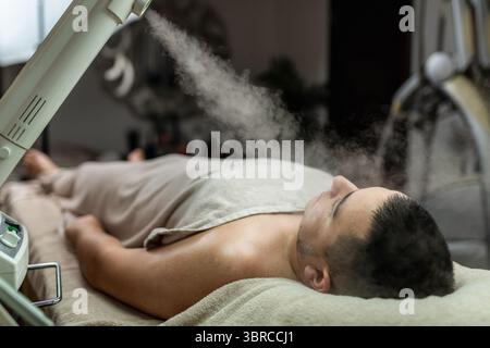 Uomo che riceve un trattamento di bellezza con un vaporizzatore viso ozono presso un centro di bellezza professionale, progettato per purificare, idratare e ringiovanire la pelle del viso per un aspetto fresco e sano. Foto Stock