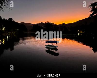 Spettacolare laguna di Huacachina con piccola barca per i turisti davanti a un bellissimo tramonto, il tutto specchiato in acqua. Lago situato in una piccola oasi. Foto Stock