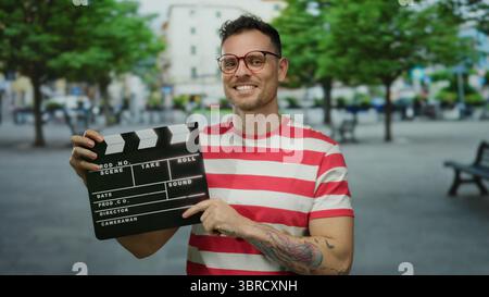 Bell'uomo sorridente sulla strada della città che tiene in mano il clapperboard con occhiali da camicia a righe e tatuaggi sullo sfondo urbano che creano vivaci ambientazioni all'aperto Foto Stock