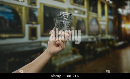 Tenendo a mano un vaso di vetro con dollari americani all'interno di un museo, illustrando temi finanziari con dipinti sfocati sullo sfondo elegante Foto Stock