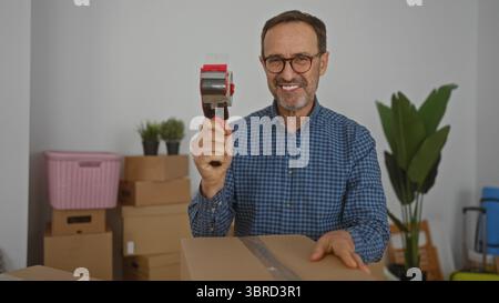 Uomo che imballa scatole di cartone in un nuovo interno del salotto con occhiali e camicia a scacchi con piante sullo sfondo che mostrano il processo di movimento. Foto Stock
