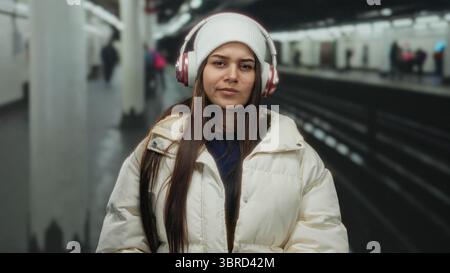 Donna che indossa le cuffie in una stazione ferroviaria al coperto, indossata in abiti invernali, in piedi su una piattaforma ferroviaria, trasudando un'espressione penosa in un bu Foto Stock