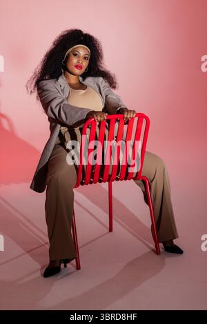 Donna afroamericana che indossa un blazer seduto all'indietro su una sedia rossa in uno studio rosa Foto Stock