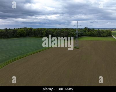 Veduta aerea di un pilone elettrico in piedi tra le texture contrastanti di campi appena lavorati e foresta verdeggiante sotto un cielo rigoglioso, quindi Foto Stock
