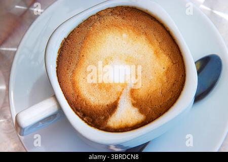 Vista ravvicinata di un cappuccino con crema di latte in una tazza quadrata di ceramica bianca, posizionata su un piattino. Vista dall'alto. Messa a fuoco selettiva Foto Stock