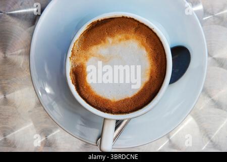 Vista dall'alto di un cappuccino con crema di latte in una tazza rotonda di ceramica bianca su un piattino servito su un tavolo metallico nel caffè all'aperto. Messa a fuoco selettiva. Foto Stock