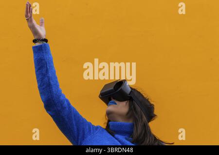 Ritratto isolato di una donna con abiti blu che indossano occhiali vr e toccano qualcosa di invisibile nella realtà virtuale. Sfondo giallo Foto Stock