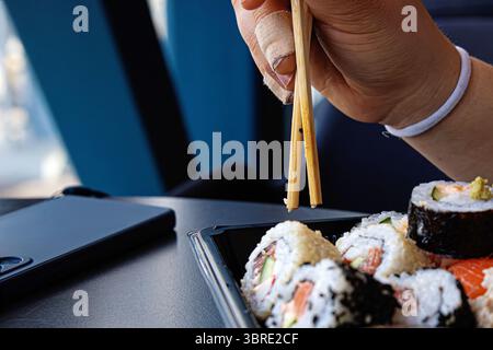 Mano con l'intonaco usando le bacchette per mangiare il sushi dal contenitore da asporto sul tavolo scuro. Cena informale con uno smartphone nelle vicinanze. Messa a fuoco selettiva Foto Stock