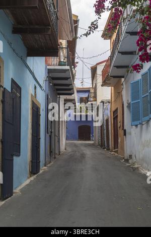 Veduta di un viale soleggiato dove si trovano edifici blu e pesca vivaci, adornati da balconi in legno e bouganville fiorite, Pano Lefkara, Larnaca, Cipro. Foto Stock