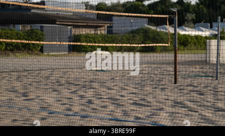 Primo piano di una rete da Beach volley che si estende su un campo di sabbia vuoto vicino a un'area di villeggiatura. La luce del sole del mattino proietta lunghe ombre, suggerendo un ea Foto Stock