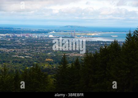 Ampia vista panoramica di Dublino, caratterizzata da un'espansione urbana, strutture industriali con camini e un'ampia baia sotto un cielo nuvoloso. Alberi sempreverdi fr Foto Stock