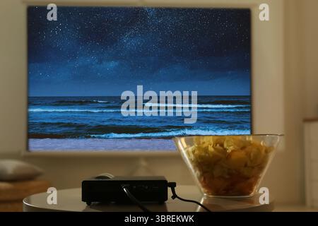 Videoproiettore e ciotola con patatine sul tavolo di legno. Vista del mare notturna sullo schermo di proiezione Foto Stock