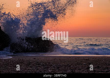 Una potente onda si schianta contro una roccia sulla spiaggia di Megali Petra, Lefkada, illuminata dalle vibranti tonalità arancio e rosa di un tramonto mediterraneo. Dyna Foto Stock