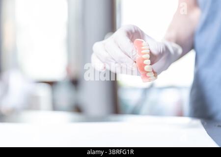 Clinica odontoiatrica luminosa con guanti da dentista che regge una dentatura superiore per pazienti anziani, concentratevi sulla dentatura, braccio offuscato Foto Stock