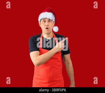 Foto in studio di un uomo che indossa un grembiule rosso su sfondo isolato. Sorpreso puntare il dito verso un lato, bocca aperta espressione stupita Foto Stock