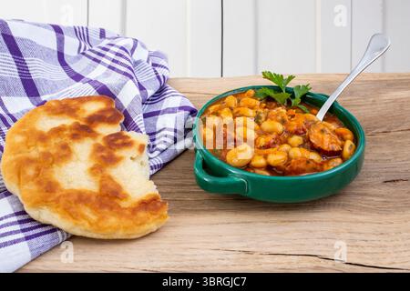 Stufato di fagioli di burro con salsa di pomodoro e salsicce di maiale a fette, pane pita e un asciugamano viola su un tavolo in legno di quercia Foto Stock
