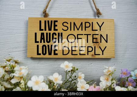 Vivi semplicemente ridi spesso ama profondamente il testo su cartelli in legno appesi e fiori decorati su sfondo bianco in mattoni Foto Stock