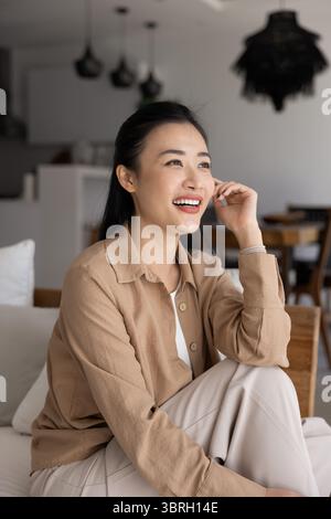 Donna seduta sul divano, che sogna ad occhi aperti, che guarda in lontananza con il sorriso Foto Stock
