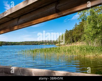 Una serena vista lago incorniciata da travi di legno, che sfoggiano lussureggiante vegetazione e acque calme sotto un cielo blu limpido. Foto Stock