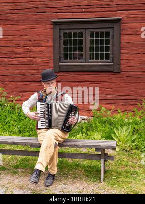 Sodertalje, Svezia - 6 giugno 2025: Musicista che suona la fisarmonica seduto su una panchina di legno di fronte ad un rustico edificio rosso. La scena è sur Foto Stock