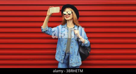 Ritratto di una giovane donna sorridente che scatta selfie con lo smartphone che indossa la giacca jean, cappello rotondo nero su sfondo rosso Foto Stock