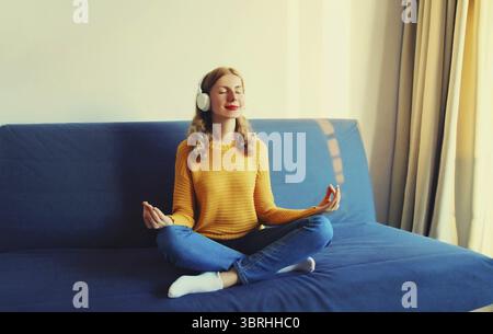 Giovane donna felice e rilassata che ascolta la musica con le cuffie wireless che meditano a occhi chiusi seduti in posizione di loto sul divano di casa Foto Stock