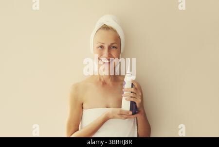 Ritratto di una donna caucasica di mezza età sorridente e felice con denti bianchi e puliti tiene l'irrigatore orale e si asciuga i capelli bagnati con un asciugamano avvolto sul cigno Foto Stock