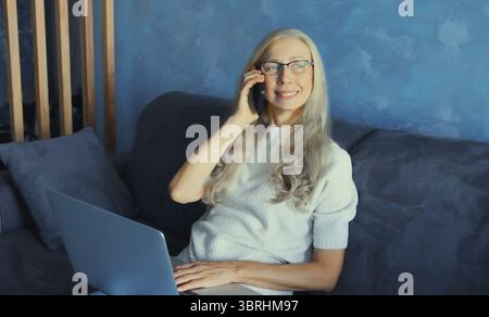 Una donna matura moderna che lavora con un laptop che parla al telefono, prende le chiamate seduto su un divano nel soggiorno di casa Foto Stock