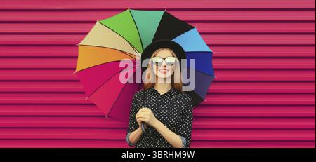 Ritratto di una giovane donna sorridente ed elegante con un ombrello colorato in cappello rotondo nero e occhiali su sfondo rosa Foto Stock