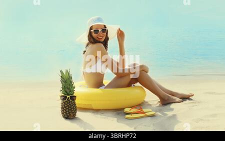 Vacanze estive, giovane donna felice e rilassante sdraiata sulla sabbia sulla spiaggia con anello gonfiabile per nuotare, ananas, occhiali da sole e infradito sul retro del mare Foto Stock