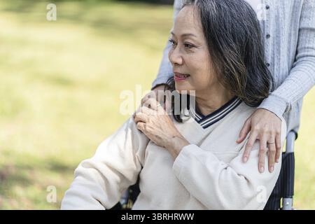 Rapporto di famiglia donna asiatica anziana in sedia a rotelle con figlia felice che tiene l'assistente per mano mentre trascorre del tempo insieme Foto Stock