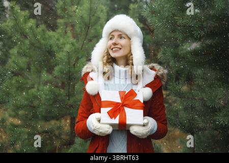 Ritratto di una giovane donna felice sorridente che tiene in mano una scatola regalo rossa festosa sullo sfondo dell'albero di Natale nella foresta invernale, con cappello bianco, Jack Foto Stock
