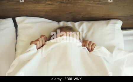 Sonno sano, donna sdraiata a letto sotto coperta bianca o piumone che nasconde il viso in camera da letto a casa Foto Stock