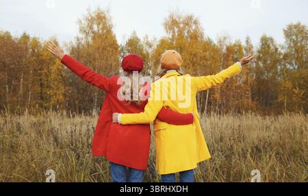 Vista sul retro, due giovani amiche felici nel parco autunnale che si godono il caldo tempo, eleganti fidanzate che alzano le mani insieme indossando il giallo rosso co Foto Stock