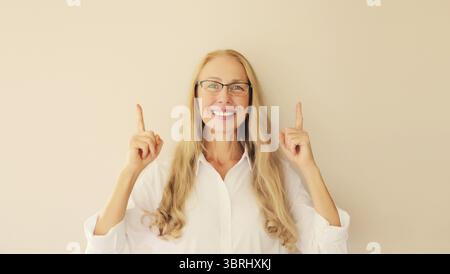 Ritratto di una donna di mezza età felice, sorridente, con gli occhiali che punta le dita verso l'alto e guarda la fotocamera sullo sfondo beige dello studio, poliziotto vuoto Foto Stock