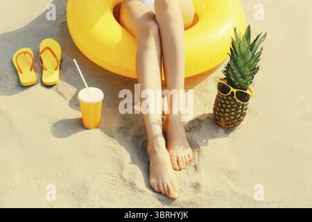 Vacanze estive, gambe da vicino di donna rilassante sdraiate sulla sabbia sulla spiaggia con anello gonfiabile giallo nuoto, ananas in occhiali da sole, tazza di juic Foto Stock