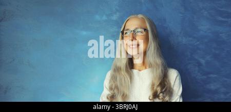 Ritratto di una donna di mezza età felice sorridente e sicura con occhiali da vista. Donna dai capelli grigi in casual su sfondo blu Foto Stock