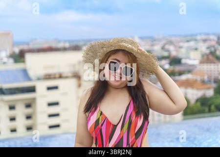 Giovane donna asiatica in sovrappeso che indossa costume da bagno arancione e cappello da spiaggia in paglia che si rilassa in piscina donna felice e di taglia allegra vacanza divertente in viaggio Foto Stock