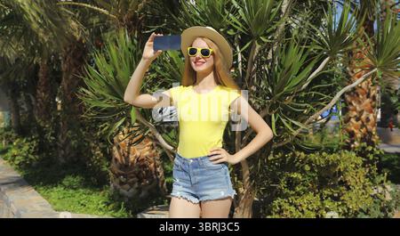 Vacanze estive, felice sorridente giovane donna che si fa selfie con il telefono in cappello di paglia da turista, occhiali gialli nel parco con palme Foto Stock