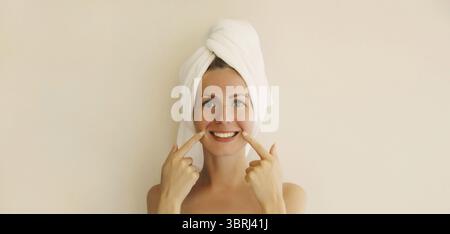 Il ritratto di una giovane donna caucasica sorridente e felice punta ai suoi denti bianchi e puliti mentre si asciuga i capelli con un asciugamano sulla testa dopo la doccia nei morni Foto Stock