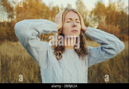 Una giovane donna stanca turbata con mal di testa, dolore alla testa, soffre di emicrania in piedi all'aperto nel parco autunnale Foto Stock