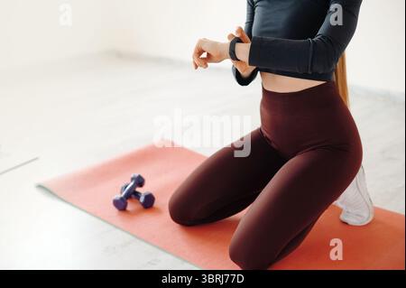 Una donna del fitness seduta sulle ginocchia su un tappetino in gomma con attrezzature sportive al chiuso. Donna slanciata vestita con leggings stretti di colore rosso scuro e maglia lunga nera Foto Stock