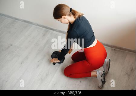 Vista superiore di una determinata donna seduta sulle ginocchia e che si esercita con manubri sul pavimento dello studio di formazione. Donna sana che esegue allenamento Foto Stock
