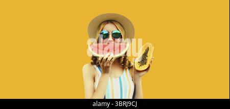 Ritratto estivo di una donna felice che mangia una fetta fresca di anguria e papaya con cappello di paglia e occhiali da sole su sfondo giallo Foto Stock