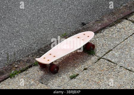Un piccolo skateboard rosa con ruote rosse e un ponte testurizzato riposa abbandonato ai margini di un marciapiede in cemento e di una strada asfaltata, evocando nostalgia Foto Stock