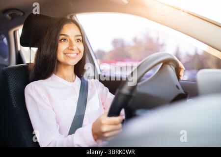 Giovane donna indiana sorridente con fiducia durante la guida dell'auto, mani sul volante Foto Stock