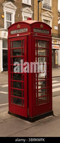 Una tradizionale cabina telefonica rossa si trova su un marciapiede di fronte ad un edificio in mattoni. Lo stand presenta un design classico con pannelli in vetro e una corona Foto Stock